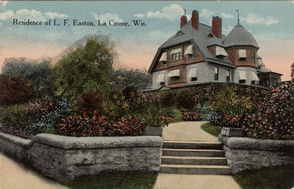 View of the three-story home, including a turret, of the L.F. Easton residence, with a stone wall, stairs leading to a front walk, and a sidewalk in the foreground. There are shrubs and plants in the front yard gardens. Caption reads: "Residence of L. F. Easton, La Crosse, Wis."