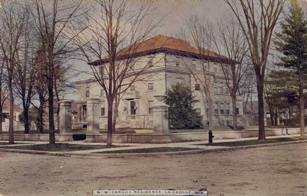 View across intersection towards the home of W.W. Cargill, founder of Cargill, a grain company. There is a low, stone wall, and columns flanking the property along the sidewalk. Caption reads: "W.W. Cargill Residence, La Crosse, Wis."