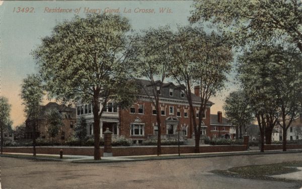 View across intersection towards the home of Henry Gund, founder of Gund Brewing. There is a low brick and stone wall, and columns flanking the property along the sidewalk. Caption reads: "Residence of Henry Gund, La Crosse, Wis."