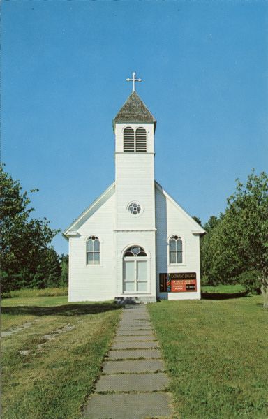 View down sidewalk towards the front of a small wooden church. Text on back reads: "Franciscan Fathers, Madeline Island."