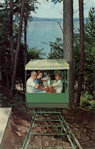 Photographic postcard view of three people on the hillside lift at Chateau Madeleine. Lake Superior is at the bottom of the hill. Text on back reads: "Summer and fall resort on Madeline Island (Apostle Islands) in Lake Superior. Less energetic guests (and others) enjoy the scenic ride in the 'hillside lift' from the Chateau to the beach and Sea Cottage."