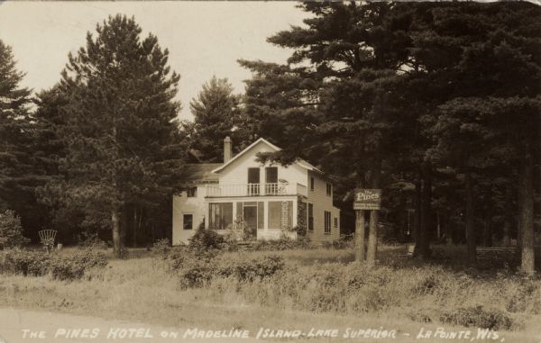 View from road towards a small wooden hotel among the pines. There is a balcony and a screened-in porch. A sign on a tree on the right reads: "The Pines, Rooms — Meals, Camping & Picnic Grounds."