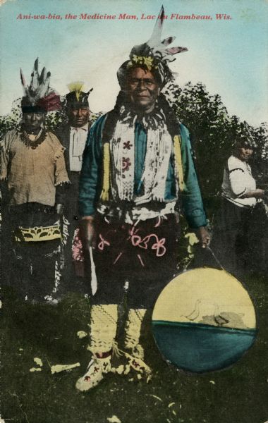 Colorized postcard of a Lac du Flambeau medicine man in traditional dress holding a drum and a knife. Other people are standing behind him. Caption reads: "Ani-wa-bia, the Medicine Man, Lac du Flambeau, Wis."