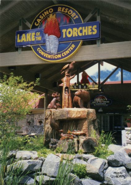 View of a wooden sculpture at the entrance of a casino. Text on reverse reads: "This carving by Gene Delcourt is representative of the seven clans of the Lake Superior Chippewa Indians and rests at the entrance to the Lake of the Torches Casino Resort in Lac du Flambeau, Wisconsin."