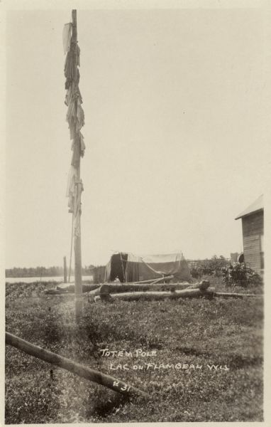 View of a pole with cloth fabric hanging on it. The side of a building is on the right. Caption reads: "Totem Pole, Lac du Flambeau, Wis."