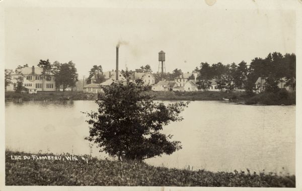 View of the village of Lac du Flambeau from across a river. Caption reads: "Lac du Flambeau, Wis."
