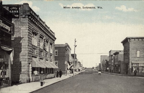 View of the businesses along Miner Avenue. A grocery is in the left foreground, with a sign for the State Bank on the brick wall of the building next door. Caption reads: "Miner Avenue, Ladysmith, Wis."