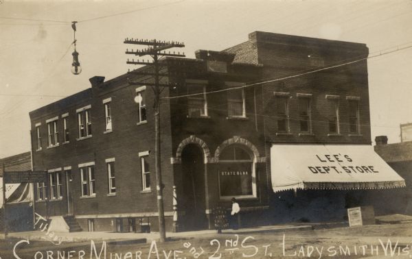 View of a central intersection and city block with the State Bank on the corner, and Lee's Department Store (Ben C. Lee & Co.) next door on the right. A sign above the sidewalk on the left reads: "Manning(?) Clark & Haviland, Real Estate Dealers, Timber Farm Lands & City Property." Caption reads: "Corner of Miner Ave. and 2nd St., Ladysmith, Wis."