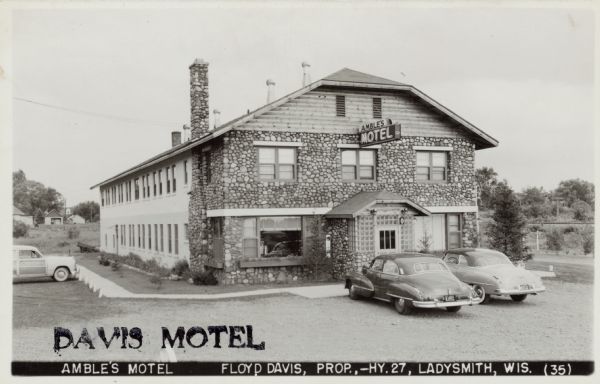 View toward the two-story motel with a stone facade. Automobiles are parked in front near the entrance, and along the left side of the building. Caption reads: "Amble's Motel, Floyd Davis, Prop — Hwy 27, Ladysmith, Wis."