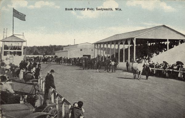 View of the race track and grandstand at the county fair. Men leading horses on the track. Caption reads: "Rusk County Fair, Ladysmith, Wis."