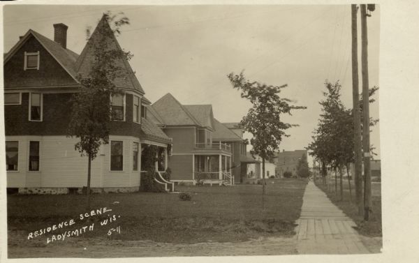 View down sidewalk of a residential neighborhood. There is a row of two-story wood-frame houses on the left, and commercial buildings are in the distance. Caption reads: "Residence Scene, Ladysmith, Wis."