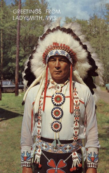 Outdoor portrait of a Native American man wearing a headdress, and bead work. Caption on back reads: "A Colorful American Indian."

