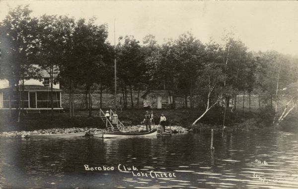 View across lake towards a building with a screened-in porch on the shore. Two men are standing on a pier with rowboats docked nearby. Two women are standing on the steps leading down to the lakefront.