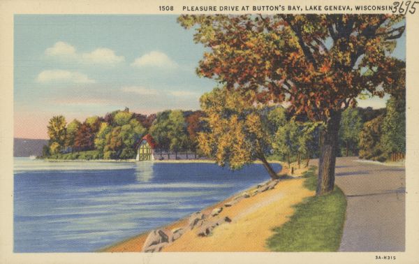 View along road and shoreline, towards a boathouse on the lake among trees with fall colored foliage. 