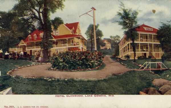 Colorized postcard view of three buildings with verandas, comprising a hotel on a hill. People are gathered on the lawn around a picnic table. A large flowering bush is planted in a circle in the center of the drive in the foreground. Caption reads: "Hotel Glenwood, Lake Geneva, Wis."