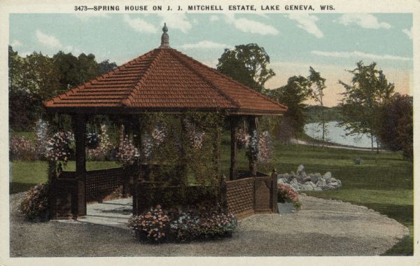 View of a gazebo with hanging flower baskets. Lake Geneva is in the background. Caption reads: "Spring House on J. J. Mitchell Estate, Lake Geneva, Wis."