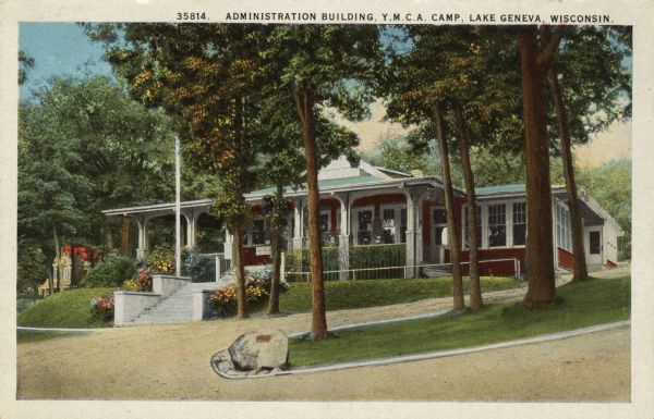 View across driveway towards a building with a front porch on a hill, with trees and flowering bushes flanking the stairs leading up to the entrance. Caption reads: "Administration Building, Y.M.C.A. Camp, Lake Geneva, Wisconsin."