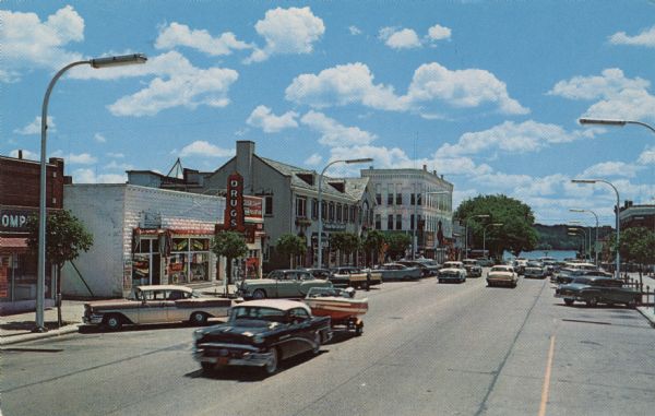 View of the central business district with the street ending at the lakefront. Automobiles are moving up and down the street, one pulling a boat on a trailer, and more cars are parked along the curbs. A drugstore is on the left.