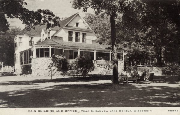 View of a wooden structure with a porch along two sides, and a flight of steps to the entrance. People are sitting and standing near benches on the shaded lawn in front. A parking lot is in the background. Caption reads: "Main Building and Office &#8212; Villa Immanuel, Lake Geneva, Wisconsin."