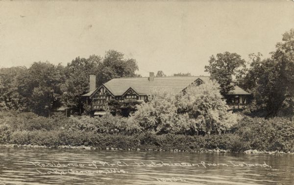 Photographic postcard view across lake towards the Tudor-style lakeside home. Caption reads: "Residence of Mr. C.L. Hutchinson from Island, Lake Geneva, Wis."