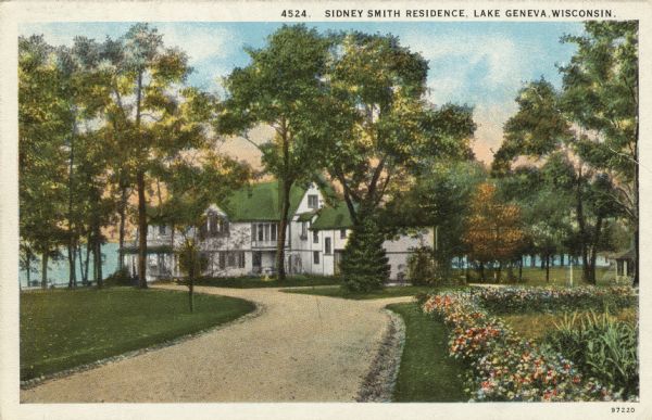 Hand-colored postcard view down driveway towards a lakeside home. Flower beds are along the driveway, and Lake Geneva is in the background. Caption reads: "Sidney Smith Residence, Lake Geneva, Wisconsin."