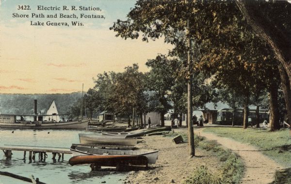 View along shore path at Lake Geneva. An electric railroad stop is behind the trees. Rowboats, canoes and an excursion boat are lined up along the beach and piers. People are standing near buildings in the background. Caption reads: "Electric R.R. Station, Shore Path, and Beach, Fontana, Lake Geneva, Wis."