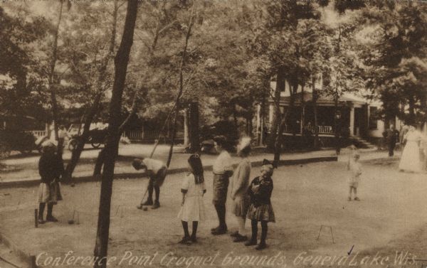 Photographic postcard view of young people playing croquet on a court surrounded by trees. Men and women are standing on the right watching. There are automobiles in the background on the left, and a large house with a porch on the right. Caption reads: "Conference Point, Croquet Grounds, Geneva Lake, Wis."