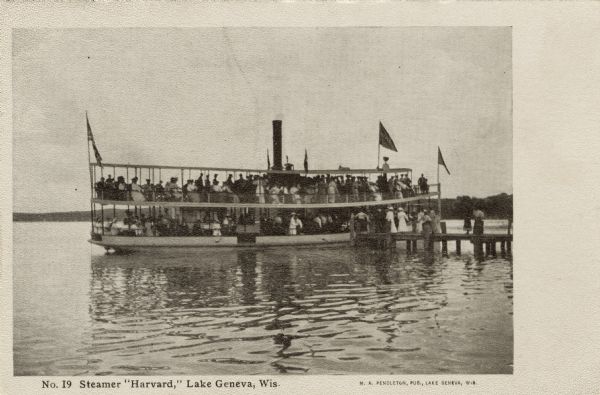 View across water towards an excursion boat filled with tourists at the end of a pier. Caption reads: "'Steamer "Harvard,' Lake Geneva, Wis."