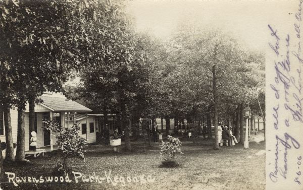 View across lawn towards cabins at a lakeside park. People are gathered on the lawn under the trees. Caption reads: "Ravenswood Park -- Kegonsa."