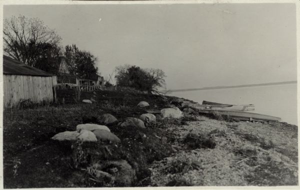 View along rocky shoreline towards a row of cottages on the shore of Lake Koshkonong. Boats are pulled up on the right, and the far shoreline is in the distance.