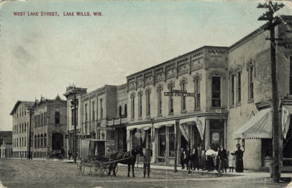 View across intersection towards a central business district, with a printer, a meat market, and a livery stable. A small crowd is gathered on the sidewalk at the corner, and a young man is standing next to a horse pulling a carriage. Caption reads: "West Lake Street, Lake Mills, Wis."