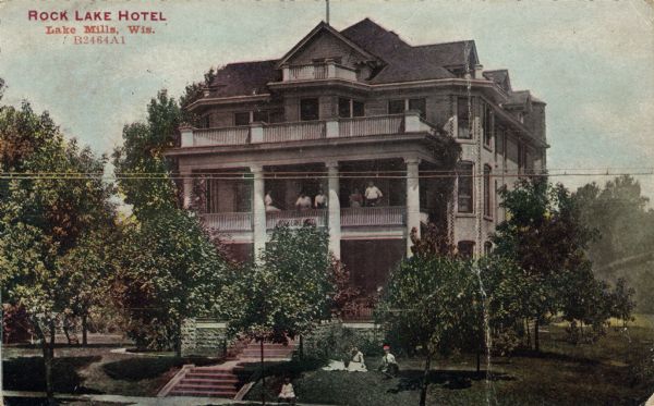 Hand-colored elevated view from across the street toward a three-story hotel with verandas on the second and third floors. People are sitting on the lawn on a hill above the sidewalk, and another group of people is standing on the second floor veranda. Caption reads: "Rock Lake Hotel, Lake Mills, Wis."