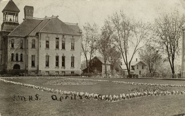 Photographic postcard view of a two-story brick school building with a bell tower. Flower beds are on the lawn, and houses and buildings are in the background. Handwritten on front: "L.M.H.S. April 22 10."