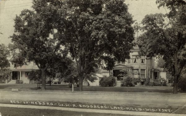 Photographic postcard view of two large Victorian homes behind trees. Caption reads: "Residence of Messrs. &#8212;  C. & W. &#8212; A. &#8212; Engsberg, Lake Mills, Wis."
