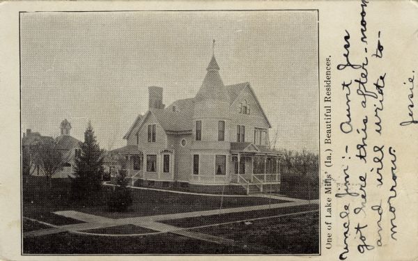 Elevated view of a corner house with a turret. Porches are on the front and side of the house, and a large stable is in the background on the left. Caption reads: "One of Lake Mills' (Ia.) Beautiful Residences, Lake Mills, Wis."
