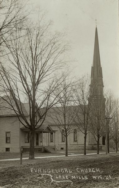 Angled view across unpaved road towards a wood-frame house, and further down the road, the Evangelical Church, both buildings facing a sidewalk. A hitching post with the shape of a horse head is along the road on the left. Caption reads: "Evangelical Church, Lake Mills, Wis."