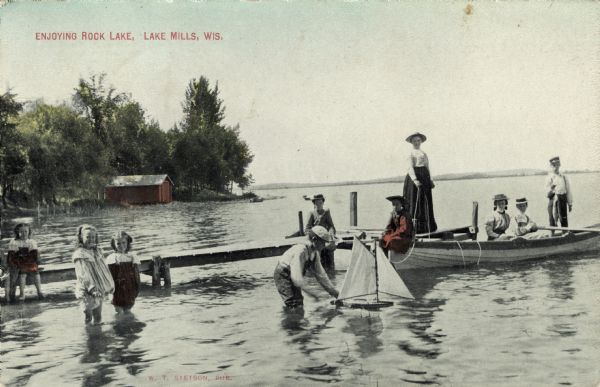 View across water towards a group of people on and next to a dock on a lake. Four people are in a rowboat. A boy is playing with a toy sailboat in the foreground. Caption reads: "Enjoying Rock Lake, Lake Mills, Wis."