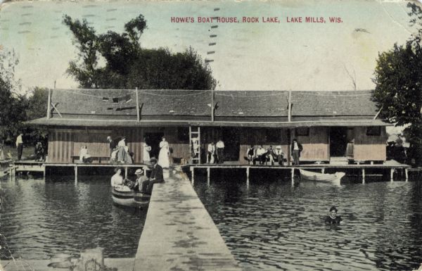 View down pier towards a boathouse on the shore, with groups of people sitting and standing on the platform in front. A group of people are in a rowboat next to the pier on the left, and a man is swimming on the right. Caption reads: "Howe's Boat House, Rock Lake, Lake Mills, Wis."