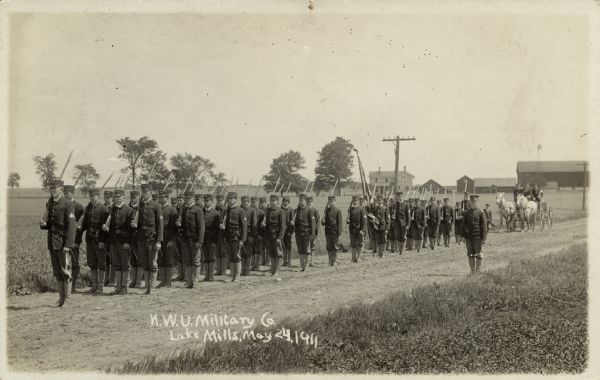 A group of soldiers with rifles standing on a dirt road. A horse and carriage are behind them, and a farm with farm buildings are in the distance. Caption reads: "N.W.U. Military Co., Lake Mills."