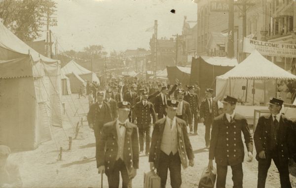 Slightly elevated view of a group of firefighters walking down a tent-lined street in downtown Lake Mills. A banner reads: "Wellcome Firefighters."