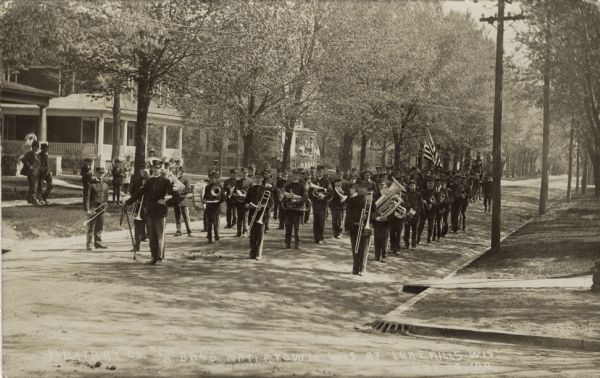 View across intersection towards a marching band carrying their instruments coming down a residential street. They are followed by a group of men in uniform carrying a flag. People are watching while standing on porches and the sidewalk. Caption at bottom reads: "Military Co. and Band, Watertown, Wis. at Lake Mills, Wis."