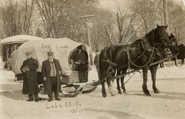 View of a group of men standing in the snow next to a covered horse-drawn sleigh. One man on the left is standing and holding a puppy in his arms; a dog is sitting next to a man in the sleigh. There is a pavilion in the background on the left. Caption reads: "Bus Line, Lake Mills, Wis."