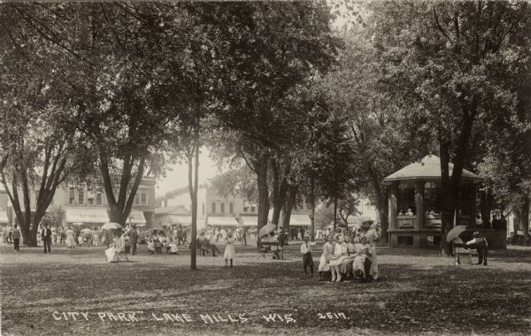 Large group of people gathered in a centrally located park, with a row of shops across the street. There is a gazebo under the trees on the right. Caption reads: "'City Park', Lake Mills, Wis."