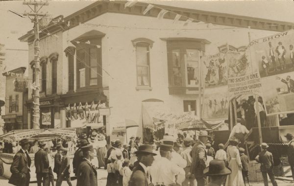 Slightly elevated view of a crowd standing in front of a stage set up in the middle of a city street, with a curtain advertising minstrel shows that reads: "Old Plantation Show" and "Two Shows in One, Strictly High Class & Instructive, for Ladies, Genteleman and Children." Street vendors are near the entrance.