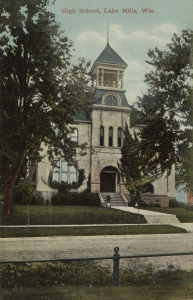 View across street towards the facade of the high school, which has an arched entrance with a bell tower above. Caption reads: "High School, Lake Mills, Wis."