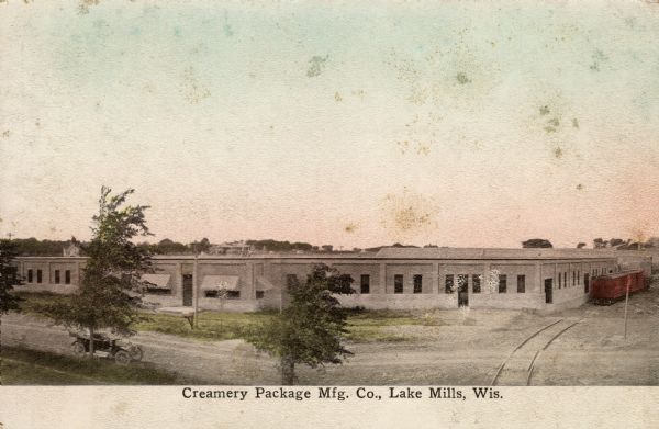 Elevated view of a creamery, with curving railroad tracks running along the right side, and two box cars near a side door. A Model T is parked on the road in front in the left foreground. Caption reads: "Creamery Package Mfg. Co., Lake Mills, Wis."