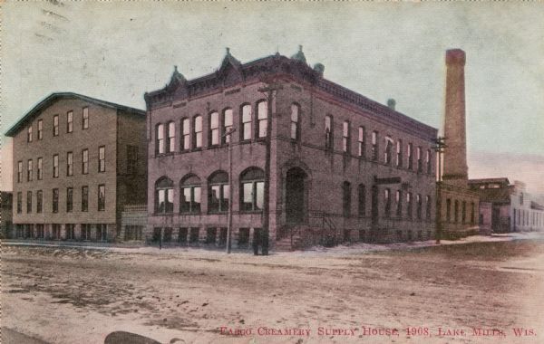 View across unpaved street towards a two-story warehouse, with a smokestack in the back. Caption reads: "Fargo Creamery Supply House, 1908, Lake Mills, Wis."