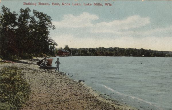 View along beach with many small stones towards three people near a beached sailboat. There is a boathouse in the background further down the shoreline. Caption reads: "Bathing Beach, East, Rock Lake, Lake Mills, Wis."