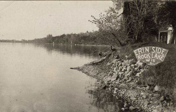 View of Rock Lake. There is a sign posted on a tree that reads: "Erin Side", and it has also been added to the postcard on the right where it reads: "Erin Side Rock Lake, Wisc." A building is in the background on the far right.