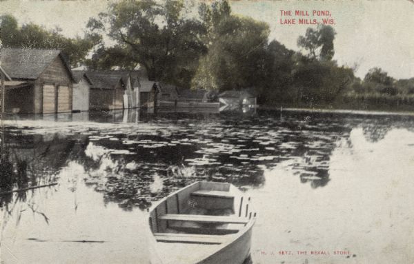 Hand-colored postcard of the Mill Pond connected to Rock Lake. A row of boathouses on the left. A small empty rowboat in the pond. Several lily pads on the surface. Caption reads: "The Mill Pond, Lake Mills, Wis."